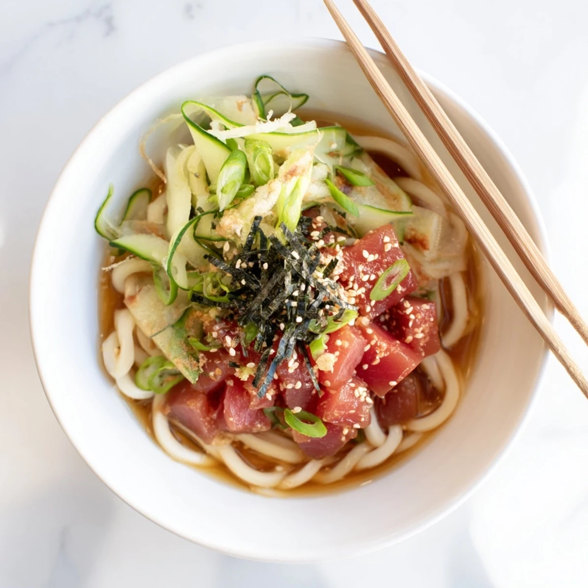 Japanese spicy tuna udon noodles served with cucumber, sesame seeds, and crispy nori in a steaming dashi broth