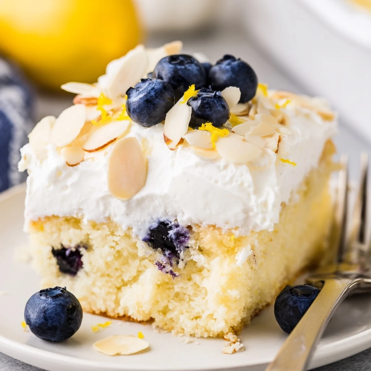 Golden lemon blueberry poke cake with creamy whipped topping and toasted almond slices garnished with fresh blueberries on a white serving plate
