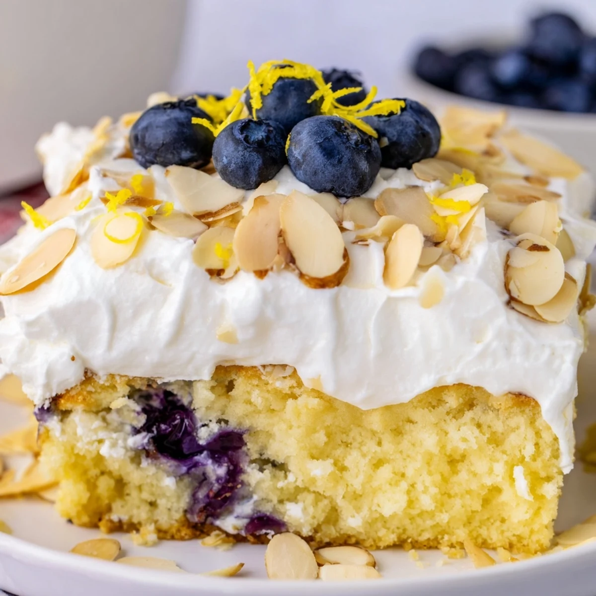Sliced lemon blueberry poke cake showing tender soaked layers whipped cream frosting and scattered toasted almonds on a rustic wooden board