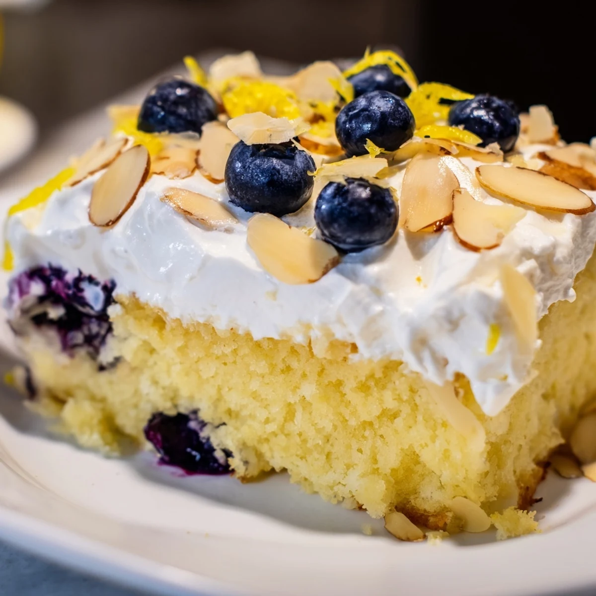 Moist lemon poke cake featuring blueberry bursts and almond crunch topped with fluffy whipped cream and lemon zest for dessert